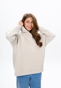 Beige oversized hoodie met een voorzak en capuchon met trekkoord, gecombineerd met blauwe spijkerbroeken, weergegeven tegen een witte achtergrond.