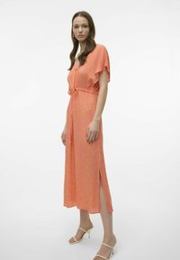Vestido largo rosa con patrón floral, mangas cortas, cintura con cordón y aberturas laterales. Combinado con sandalias de tacón beige.