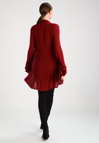Roter, langärmeliger Cardigan mit plissiertem Design, der bis zur Mitte des Oberschenkels reicht, kombiniert mit schwarzen, eng anliegenden Hosen und Stiefeletten.