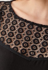 Vêtement noir avec un col en dentelle orné de motifs floraux circulaires, complété par un tissu noir lisse en dessous. Bords festonnés détaillés.