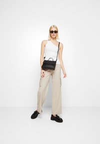 Black shoulder bag with a top handle, white asymmetrical tank top, beige cargo pants, and black chunky shoes, model standing on a plain background.