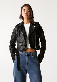 Giacca cropped in pelle nera con dettagli in argento, chiusura lampo frontale, tasche laterali e vita con cintura, abbinata a jeans blu a vita alta.