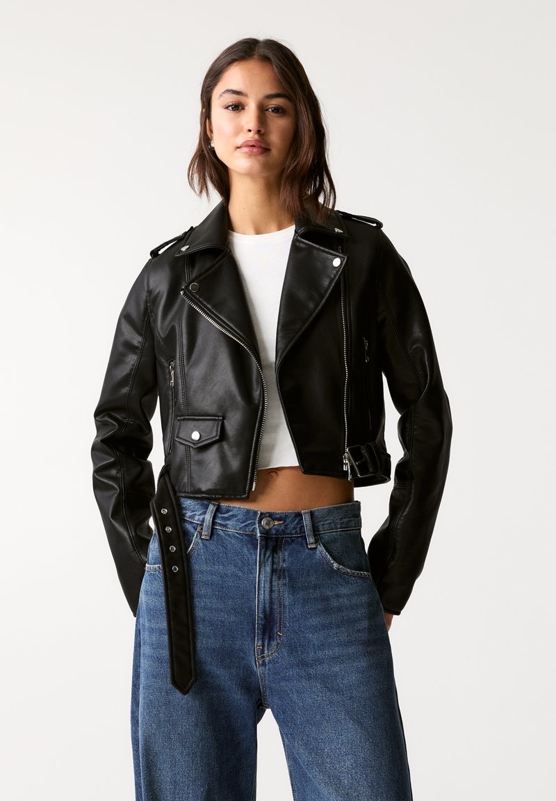 Giacca cropped in pelle nera con dettagli in argento, chiusura lampo frontale, tasche laterali e vita con cintura, abbinata a jeans blu a vita alta.