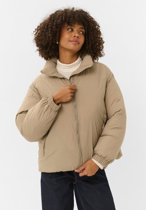 Mujer con cabello rizado que lleva una chaqueta acolchada beige y un cuello alto blanco, abrochándose la cremallera de la chaqueta, de pie frente a un fondo claro y liso.