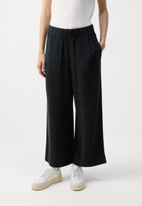 Schwarze, weit geschnittene Hosen mit elastischem Bund, aus einem glatten Stoff, kombiniert mit weißen Turnschuhen. Verfügt über seitliche Taschen.