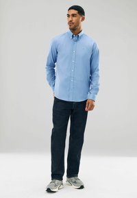 Šviesiai mėlynas marškinėlių su apykakle modelis, derinamas su tamsiais džinsais. Marškiniai turi logotipą ant krūtinės ir ilgomis rankovėmis.