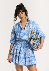 Robe à motif floral bleu avec jupe à volants, manches longues et taille à cordon. Petit sac à main en osier avec des accents bleus et naturels.