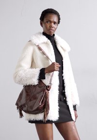 Manteau en fausse fourrure blanche avec un col châle, associé à un pull noir ajusté et un sac en cuir marron foncé avec des franges. Le modèle se tient avec confiance.