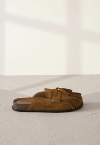 Zapato de ante marrón tipo slip-on con un diseño con flecos, forma estructurada y una suela plana negra con una textura sutil y detalles de costura.