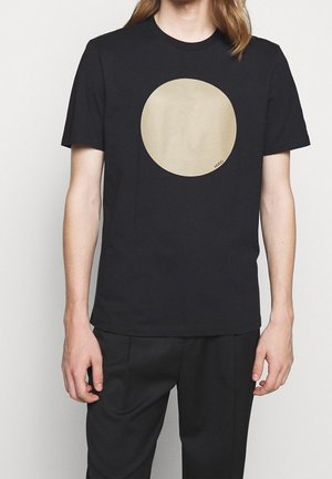Personne portant un t-shirt noir à manches courtes avec un grand cercle beige et un petit texte "HUGO" au centre devant.