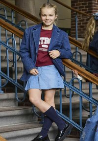 Blazer di jeans blu sopra un sudario bordeaux, abbinato a una gonna di jeans azzurro chiaro. Calze scure oltre il ginocchio e scarpe nere. Ambientazione naturale.