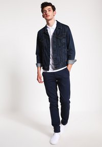 Giacca di jeans blu scuro indossata sopra una polo bianca, abbinata a pantaloni navy e sneakers bianche. La giacca ha un design classico con tasche con bottoni.