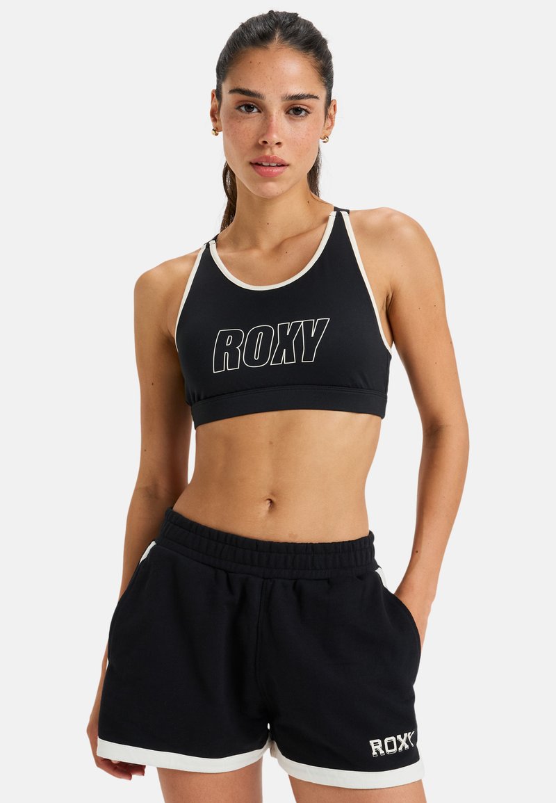 Musta urheilutoppi valkoisilla yksityiskohdilla ja edessä "ROXY"-logo, yhdistettynä mustiin shortseihin, joissa on valkoisia yksityiskohtia ja joustava vyötärö.
