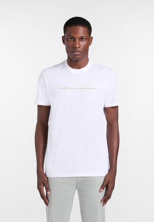 Jonge man, frontaal gezicht, draagt een wit T-shirt met korte mouwen met een dunne horizontale beige streep en tekst over de borst, gecombineerd met lichtgrijze broek.
