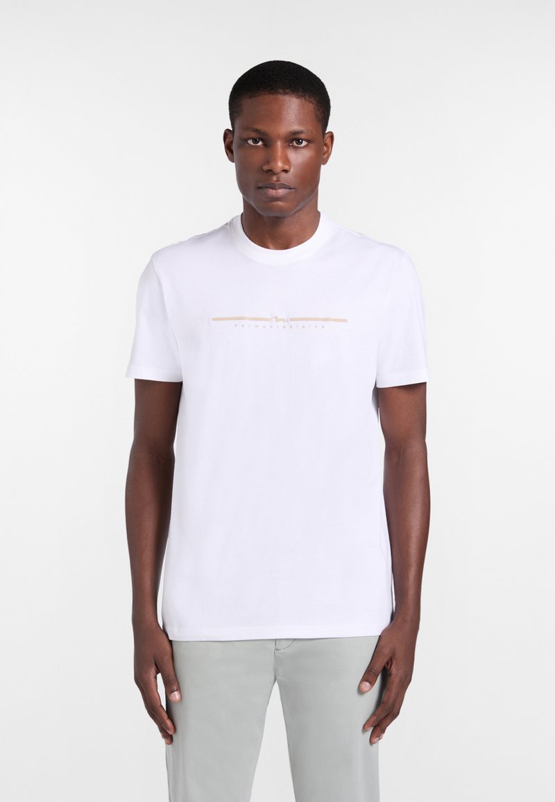 Joven de frente, vistiendo una camiseta blanca de manga corta con una delgada línea horizontal beige y texto en el pecho, combinada con pantalones gris claro.