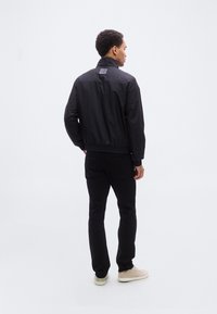 Μαύρο μπουφάν τύπου bomber με γιακά, ελαστική μέση και μανσέτες; φορεμένο με μαύρο παντελόνι και ανοιχτόχρωμα αθλητικά παπούτσια, σε όψη από πίσω.