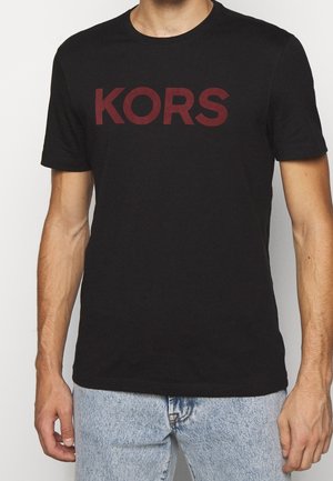 Homme portant un t-shirt noir à manches courtes avec un grand texte rouge "KORS", assorti à un jean en denim bleu clair, debout devant un fond blanc.