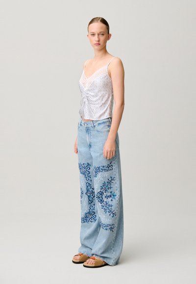 Donna con top bianco senza maniche rifinito con pizzo, jeans azzurri a gamba larga con motivi blu e sandali color cuoio, in piedi davanti a uno sfondo semplice.