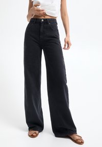 Jeans de cintura alta en denim negro de pierna ancha. Cuenta con un cierre de botón en la parte delantera, trabillas para el cinturón y costuras visibles. Presenta un acabado liso y texturizado.