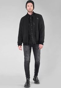 Chaqueta bomber de forro polar negra con puños acanalados, superpuesta sobre una camisa oscura abotonada y vaqueros pitillo negros, complementada con botines negros.