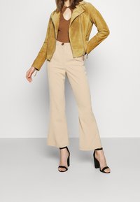 Tan suede moto jacket, brown ribbed top, beige flared trousers, and black heeled sandals showcase a layered, monochromatic ensemble.