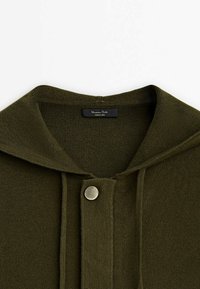 Cardigan en maille vert foncé avec un large col et un seul bouton-pression rond en argent en haut au centre.