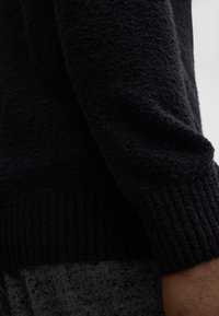 Dunkles Sweatshirt mit einer strukturierten, plüschigen Oberfläche und gerippten Bündchen, kombiniert mit einem grauen Stoff im Fischgrätmuster darunter.