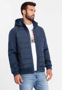 Mörkblå hybridjacka med vadderad framsida och huva, tillverkad av slätt tyg. Har ribbade ärmslut, sidofickor och dragkedja framtill.