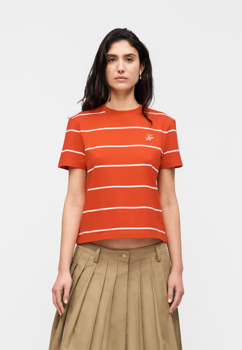 Frau mit langen dunklen Haaren, die ein oranges kurzärmliges Shirt mit weißen horizontalen Streifen und einen beigen plissierten Rock trägt.