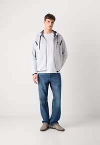 Haut de sweatshirt zippé en polaire gris clair avec des détails en mesh, accents noirs, poches kangourou, porté sur un t-shirt blanc et un jean bleu, associé avec des chaussures beiges.