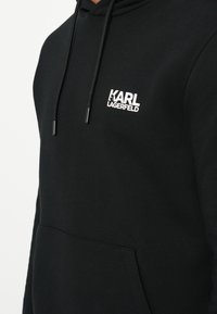 Čierna mikina s kapucňou a prednou kapsou, na ľavej hrudi s bielym logom "KARL LAGERFELD" a čiernymi šnúrkami na kapucni.