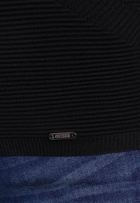 Zwarte gebreide trui met een ribbeltextuur en een klein zilveren logo-opdruk aan de zoom. Gecombineerd met denimstof eronder.