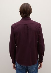 Bordeauxrotes Langarmhemd mit spitzem Kragen, Rückenfalte und geknöpften Manschetten, kombiniert mit blauen Denim-Jeans. Glatte Stofftextur.