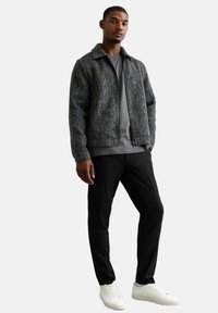 Chaqueta gris texturizada con cuello, llevada sobre una camisa gris, combinada con pantalones negros y zapatillas blancas. Diseño simple, paleta de colores neutros.