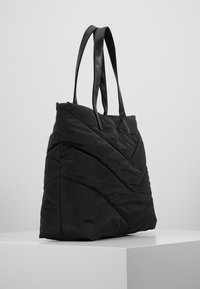 Sac en tote matelassé noir avec un coussin de couture diagonal et deux longues poignées lisses, posé sur une surface blanche contre un fond gris clair.