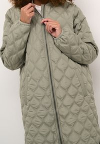 Quiltad olivgrön jacka med diamantmönster, framdragkedja och sidofickor. Tillverkad av ett mjukt, vadderat material.