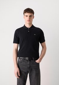 Black polo shirt with short sleeves, two-button collar, and small logo accent. Worn with dark gray jeans and a black belt.