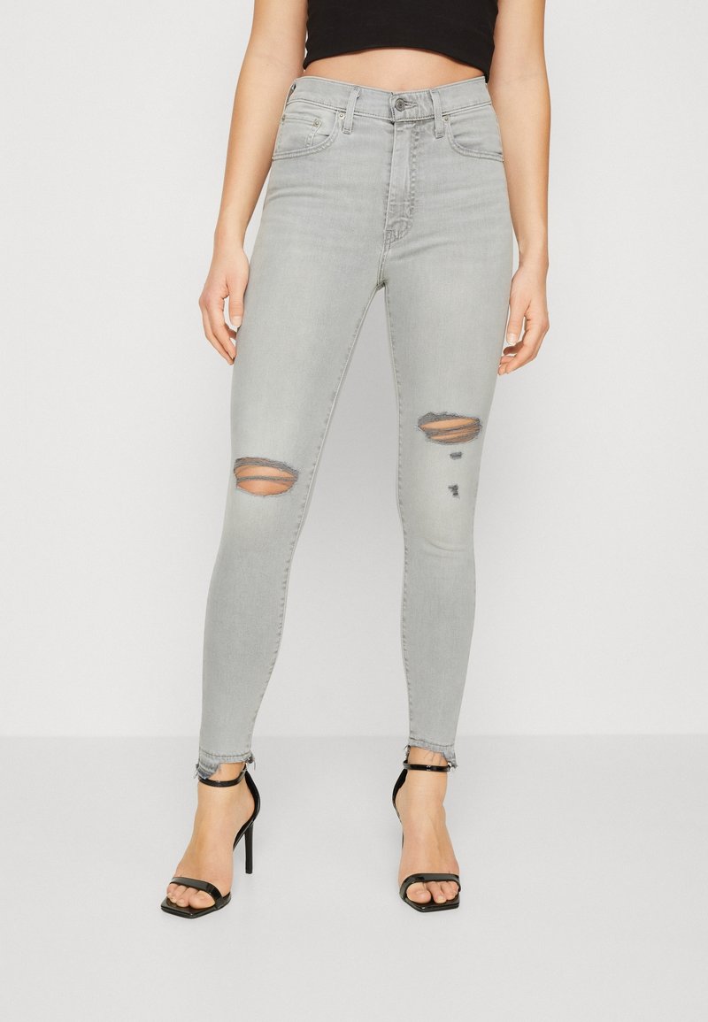 Femme portant un jean skinny taille haute déchiré gris clair et des sandales à talons hauts noires sur un fond uni.