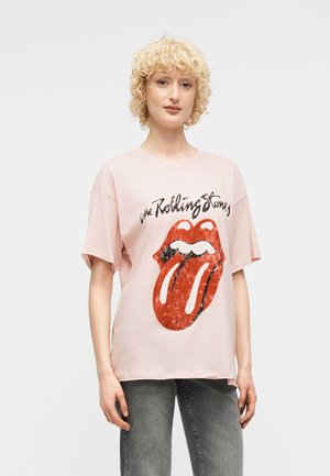 Νεαρό άτομο με σγουρά ξανθά μαλλιά που φοράει ανοιχτό ροζ μπλούζα Rolling Stones με λογότυπο, στέκεται μπροστά σε ένα απλό λευκό φόντο.
