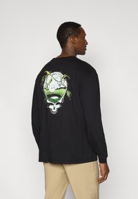 Μαύρο μακρυμάνικο μπλουζάκι με πολύχρωμο γραφικό κρανίου στην πλάτη, με πράσινα κλήματα και ένα τοπίο μέσα στο σχέδιο του κρανίου.