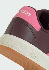 Zapatilla de cuero marrón con un acento rosa cerca del talón, suela más clara texturizada y logo de la marca embosado. Diseño simple con materiales duraderos.