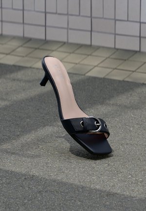 Black heeled mule sandal with square toe, silver buckle on strap, placed on asphalt near tiled wall.