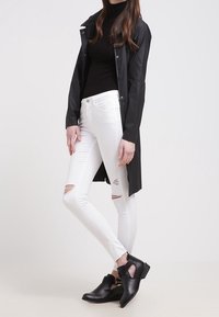 Svart vattentät kappa över en svart polotröja, vita rippade tajta jeans och svarta ankelstövlar med spänndetaljer.