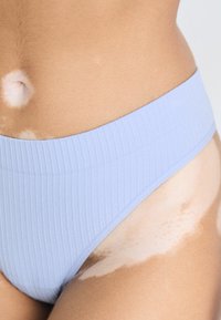 Bas de bikini à taille haute en bleu clair côtelé. Texture douce et bords lisses. Exposition notable de la peau sur les côtés, montrant des teintes de peau variées.