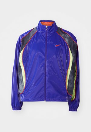 Chaqueta de nailon púrpura con un alto cuello, que presenta paneles negros y verdes, detalles rojos y puños elásticos. Cierre con cremallera en la parte delantera.