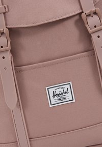 Gros plan sur un sac à dos en tissu mauve avec des boucles en plastique et une étiquette blanche de la marque Herschel sur la poche avant.