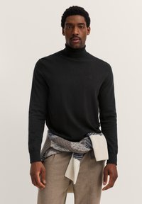 Schwarzer Rollkragenpullover aus Strickmaterial, mit langen Ärmeln und glatter Textur, kombiniert mit einem gemusterten Kleidungsstück, das um die Taille gebunden ist.