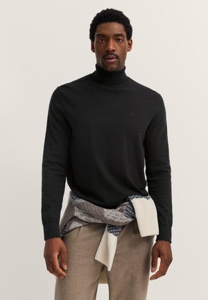 Schwarzer Rollkragenpullover aus Strickmaterial, mit langen Ärmeln und glatter Textur, kombiniert mit einem gemusterten Kleidungsstück, das um die Taille gebunden ist.