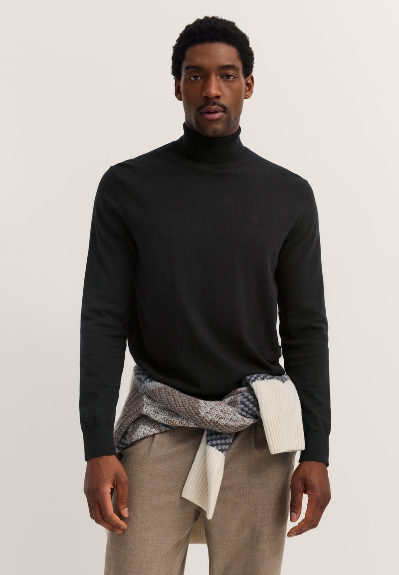 Schwarzer Rollkragenpullover aus Strickmaterial, mit langen Ärmeln und glatter Textur, kombiniert mit einem gemusterten Kleidungsstück, das um die Taille gebunden ist.