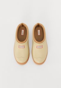 Beige gumi slip-on cipők barna talppal, lekerekített orral és saroknál húzófüllel. Piros és fehér "HUNTER" logó az oldalán.